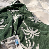 A green and white palm leaf design beach towel is laid out on the sand at the beach.