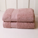 Two folded dusty pink cotton towels stacked on a white surface against a white paneled background.