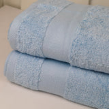 A stack of two folded light blue towels with a textured terry cloth surface and a smooth border band, photographed on a light neutral surface.