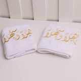 Two folded white towels with gold Arabic calligraphy embroidered on them, placed on a light surface in front of a white paneled background.