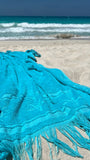 Turquoise towel with fringe on a sandy beach with ocean waves in the background